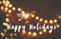 Happy Holidays text sign on glowing sparkler in hand on background of golden christmas tree lights in dark festive room. Fireworks Royalty Free Stock Photo