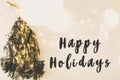 Happy Holidays text sign on modern christmas tree made of pine branches with golden festive lights hanging on white wooden wall. Royalty Free Stock Photo