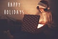 Happy Holidays text sign on stylish happy girl in santa hat opening christmas gift box with magic light in dark room. Merry Royalty Free Stock Photo