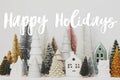 Happy Holidays text sign on stylish little Christmas trees and houses decorations on white table. Modern christmas scene, Royalty Free Stock Photo