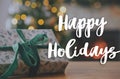 Happy Holidays text on stylish christmas gift with green ribbon close up on background of xmas tree lights and fireplace. Royalty Free Stock Photo