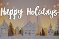 Happy Holidays text on stylish little white houses and trees, snowy christmas miniature village with golden lights. Merry Royalty Free Stock Photo