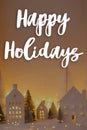 Happy Holidays text on stylish little white houses and trees, snowy christmas miniature village with golden lights. Merry Royalty Free Stock Photo