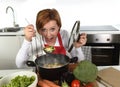 Happy home cook woman in red apron at domestic kitchen holding saucepan with soup tasting delicious vegetable stew