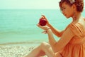 Happy honeymoon concept. Young girl in trendy vintage dress sitting on the beach and holding a red apple - love symbol. Sunny