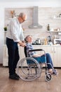 Happy husband pushing invalid senior woman in wheelchair in kitchen