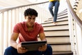 Happy ibracial man sitting on stairs at home using tablet, with legs of woman walking downstairs