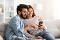 Happy indian couple sitting on sofa at home, using smartphone Royalty Free Stock Photo