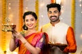 Happy indian couple in traditional ethnic wear lighting brass hanging oil lamp by looking at camera at home - concept of