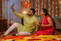 Happy Indian couples taking selfie on mobile phone during diwali festival celebration in front of rangoli at home -