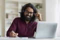 Happy indian man with headset taking notes and using laptop at home Royalty Free Stock Photo