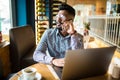 Happy indian man working at cafe with laptop. Young smiling man talking on the mobile phone while sitting at cafe and drink tea Royalty Free Stock Photo