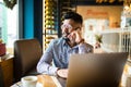 Happy indian man working at cafe with laptop. Young smiling man talking on the mobile phone while sitting at cafe and drink tea Royalty Free Stock Photo