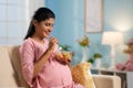 Happy indian pregnant woman eating fruit salad while sitting on sofa at home - concept of Pregnancy nutrition, Healthy