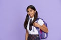 Happy indian school girl wearing uniform holding backpack on lilac background.