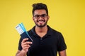 Happy indian student male bearded wearing glasses and black t-short holding passport airline ticket yellow studio