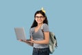 Happy Indian teen girl with backpack and laptop computer participating in online lesson on blue studio background Royalty Free Stock Photo