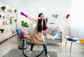 Happy Indian teen girl cleaning her home, wearing headphones, using vacuum cleaner as microphone and having fun