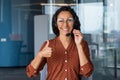 Happy indian woman in headset showing thumb up in office. Satisfied curly young businesswoman in glasses is happy to Royalty Free Stock Photo