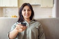 Happy indian woman sitting on sofa holding remote control watching tv at home. Royalty Free Stock Photo