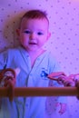 Happy infant baby is playing with wooden hanging toys in the evening in bed. Smiling child in turquoise clothes in the light of a