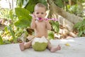 Happy infant baby is teething and gnaw a spoon. Eats and drinks green young coconut. Sits in the jungles Royalty Free Stock Photo