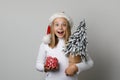 Happy joyful excited child girl in Santa hat with Xmas tree and red gift on white background. Christmas concept Royalty Free Stock Photo