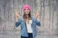 Happy joyful smiling hipster girl in casual clothes and pink hat standing against gray wall and showing victory sign, v-sign, gest Royalty Free Stock Photo