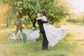 Happy just married couple dancing on lawn in green sunny park. Bicycle near decorated tree at background Royalty Free Stock Photo