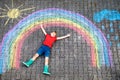 Happy kid boy in rubber boots with rainbow sun and clouds with rain drops painted with colorful chalks on ground or Royalty Free Stock Photo