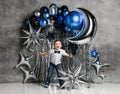 Happy kid boy stands with his hands spread wide on background with silver and blue metallic balloons for birthday party Royalty Free Stock Photo