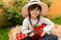 Happy kid girl in straw hat with ukulele having fun on summer field. Royalty Free Stock Photo