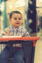 A happy kid in a shirt sits in a trolley in a shopping center and eats bread. toned