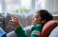 Happy kid wearing earphones listening to music on tablet, Cute boy lying on sofa playing games online on internet in living room, Royalty Free Stock Photo