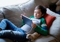 Happy kid wearing earphones listening to music on tablet, Cute boy lying on sofa playing games online on internet in living room, Royalty Free Stock Photo