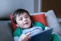 Happy kid wearing earphones listening to music on tablet, Cute boy lying on sofa playing games online on internet in living room, Royalty Free Stock Photo