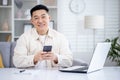 A happy Korean man is sitting in a bright living room at a table in front of a laptop, holding a phone in his hands Royalty Free Stock Photo