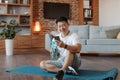 Happy korean mature man in sportswear sitting on yoga mat at home, holding bottle of water and using cellphone Royalty Free Stock Photo