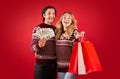 Happy lady and her boyfriend in Christmas sweaters holding fan of money and shopping bags on red background