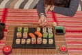 Happy lady holds a sushi roll on the chopsticks, looks at delicious Japanese food and smiles, over a table sit on a sofa Royalty Free Stock Photo