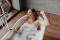 Happy lady singing song holding body brush as microphone, resting in foamy bathtub in morning