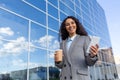 Happy Latin American business woman walks past the glass office building, holds a cup of coffee on the phone, smiles and Royalty Free Stock Photo