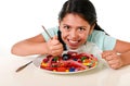 Happy Latin female child eating dish full of candy and gummies with fork and knife and big cola bottle in sugar abuse