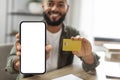 Happy latin male shopaholic showing credit card and smartphone with white blank screen, selective focus, mockup