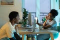 Happy African American woman showing mobile phone screen man to friend sits at table with laptop Royalty Free Stock Photo