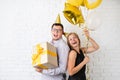 Happy laughing couple celebrating birthday party holding golden balloons and gift box over white brick wall Royalty Free Stock Photo