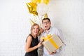 Happy laughing couple celebrating birthday party holding golden balloons and gift box over white brick wall Royalty Free Stock Photo