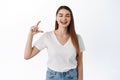 Happy laughing girl showing small size, little hand gesture and smiling, standing joyful against white background in Royalty Free Stock Photo