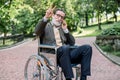 Happy laughing senior handicapped man in a wheelchair, walking in a park, using his phone, talking with friend, posing Royalty Free Stock Photo