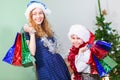 Happy and laughing sisters with color paper bags standing close to Christmas tree, kids wearing Santa hats